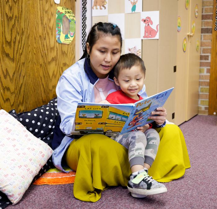An adult student who is a mother sits with her preschool aged son in her lap and reads together.
