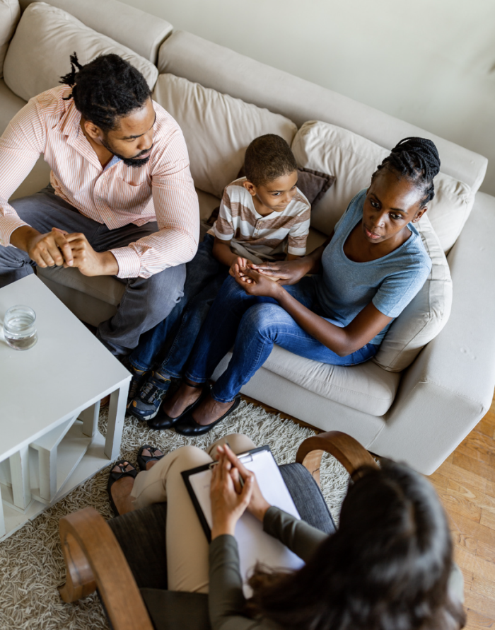 A man and a woman sit with a young boy on a couch. They are listening to information from a professional with a clip board.