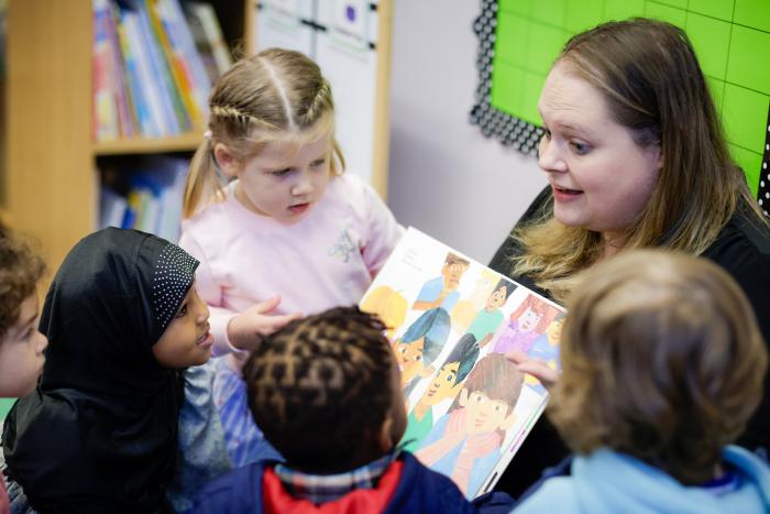 Preschool teacher reads a book to 5 preschoolers huddled around her.