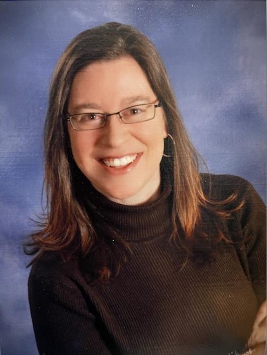 Photo portrait of Alisha smiling at the camera wearing a black turtle neck and glasses. She has mid length brunette hair.