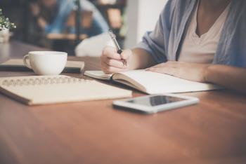 Woman writing in a notebook with a teacup and tablet beside her