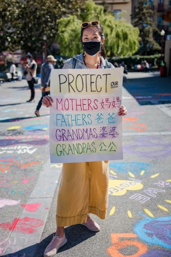 Person in a mask holding a sign reading protect our mothers fathers grandmas and grandpas