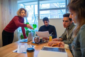 a group of four diverse adults stand and sit around the a table talking, looking at books and computers